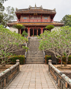 Traditional Minh Pavilion architecture at the historic Minh Mang Tomb in Hue, Vietnam, featuring ornate woodwork, a yellow tiled roof, stone stairs, and lush blooming trees in a quiet garden setting