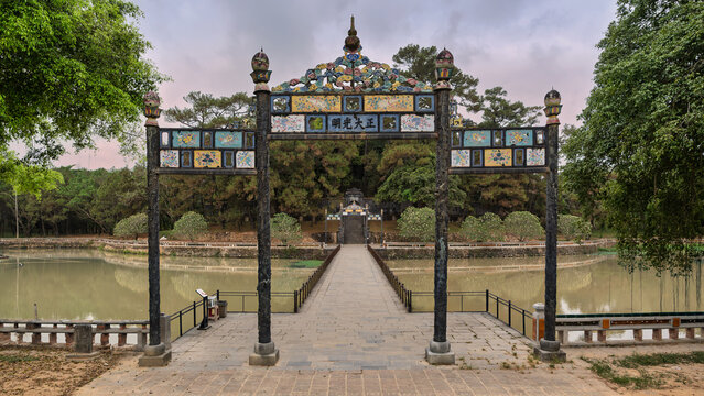 Ornate traditional gate and stone bridge leading to the Minh Mang Tomb, a historic royal burial site in Hue, Vietnam, surrounded by lush gardens and a pond