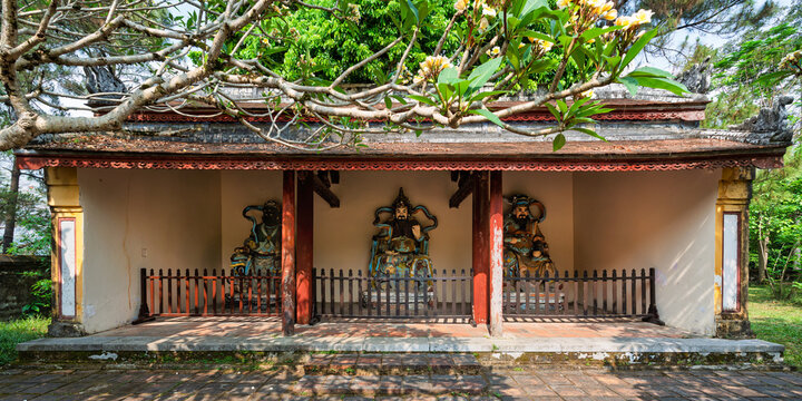 Traditional statues of Buddhist guardians in a pavilion at Thien Mu Pagoda in Hue, Vietnam, framed by blooming frangipani branches in a scenic temple setting