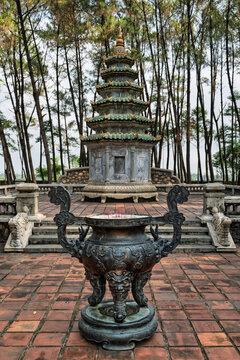 Traditional bronze incense burner with burning sticks in front of an ancient stone stupa at Thien Mu Pagoda in Hue, Vietnam, amidst a tranquil pine forest setting