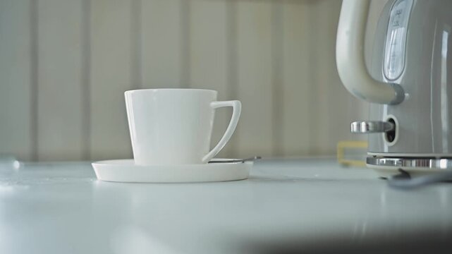 A white coffee cup rests on a saucer on a kitchen countertop near a coffee machine. The setup showcases the cup's placement and the appliance in the background