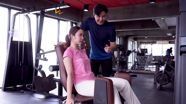 Asian fitness trainer guiding young woman through arm exercise machine session in modern gym