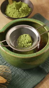 Bamboo scoop chashaku placing mound of bright green matcha powder onto metal mesh sieve over green ceramic bowl chawan on wooden table. First step of traditional matcha tea preparation.