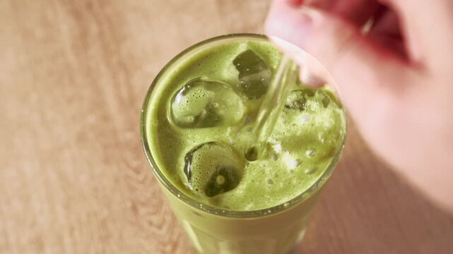 Stirring cold iced matcha green tea latte with glass straw in tall glass filled with ice cubes on wooden table. Iced matcha latte, healthy detox superfood beverage for daily summer wellness routine.