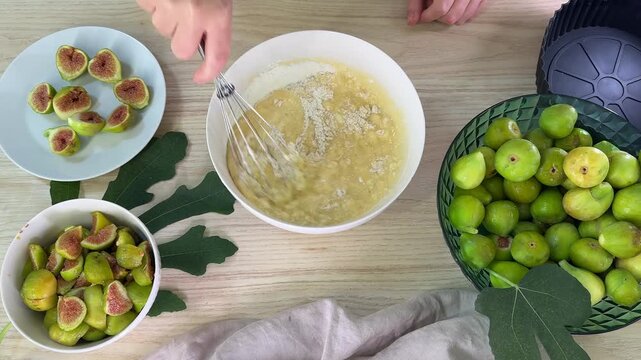 A person uses a metal whisk to stir pale yellow batter in a white bowl, surrounded by sliced and whole green figs on plates and in a basket