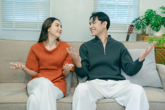 Happy Asian couple sitting on sofa talking and gesturing together at home