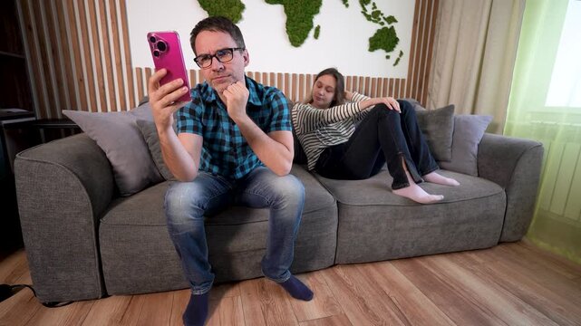 Young married couple on sofa: family conflict, silence and resentment. Husband stares at wife's phone in disbelief, or ignores her while scrolling.