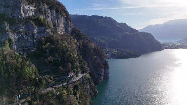 Aerial drone video of a Swiss alpine lake bordered by forested cliffs and rocky mountain slopes, with bright water reflections and distant valley views.