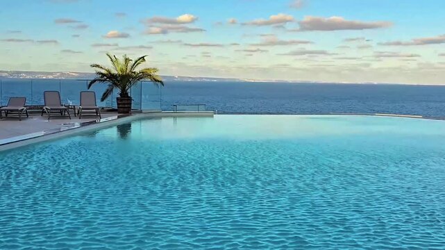 Rooftop swimming pool overlooking the sea, surrounded by palm trees, lounge chairs and glass safety railings under a blue summer sky