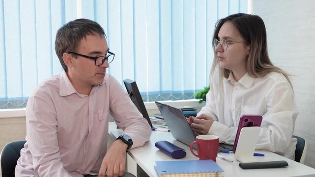 Two office colleagues, a man and a woman, talking to each other at a desk. Business communication, professional discussion, and teamwork.