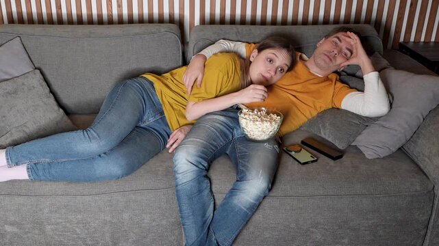 Young couple enjoying a romantic movie night on the sofa at home, watching an interesting film in the living room and sharing popcorn during a cozy date.