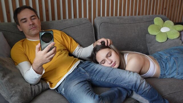 Man scrolling smartphone on sofa while tired girlfriend sleeps on his lap. Romantic relationship, lazy home leisure, and boring routine together.