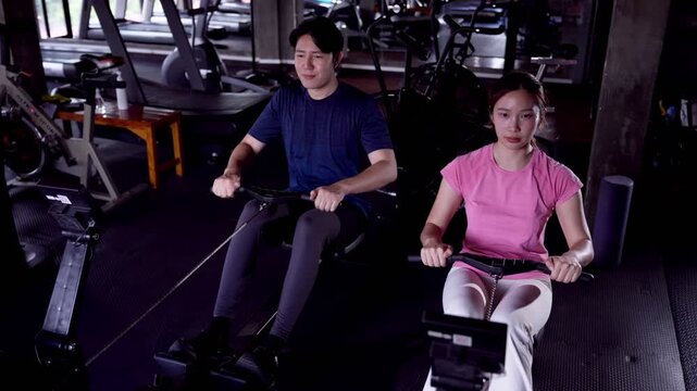 Asian man and woman using rowing machines together during intense workout in gym