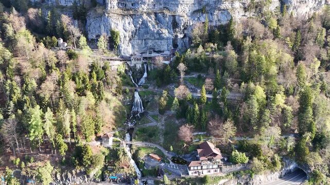 Aerial drone video of a Swiss alpine lake bordered by forested cliffs and rocky mountain slopes, with bright water reflections and distant valley views.