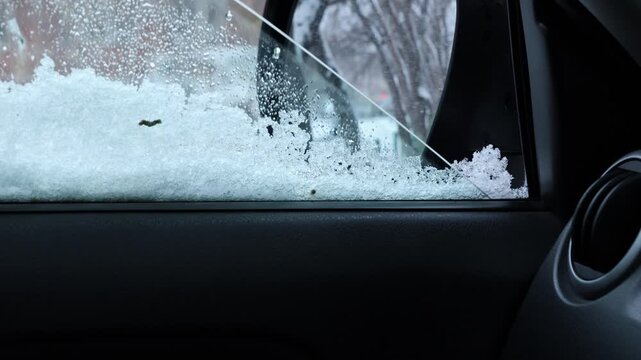 Car window partially opened as snowflakes accumulate on the glass and side mirror. Snowfall creates a wintry atmosphere outside the vehicle, with cold and icy conditions visible on the window surface.