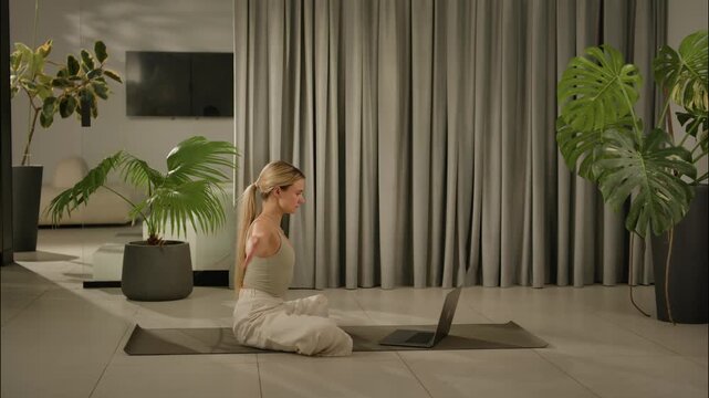 Seated yoga stretch beside laptop in calm modern home interior with tropical plants and soft curtain