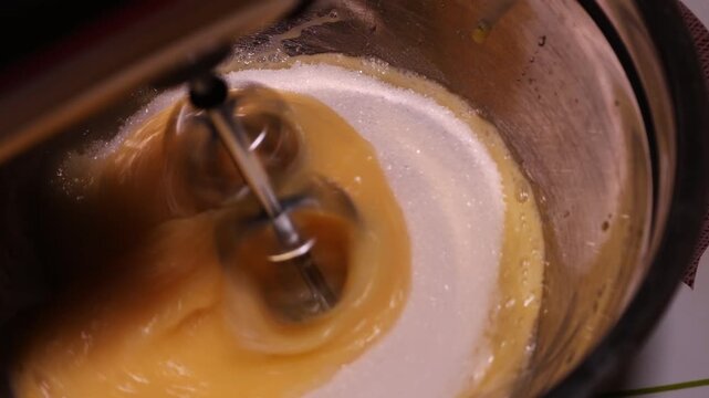 Close-up of hand mixer beating eggs combined with sugar in a metal bowl. Eggs and sugar blend into a light, airy mixture perfect for baking or cooking recipes that require a fluffy texture.