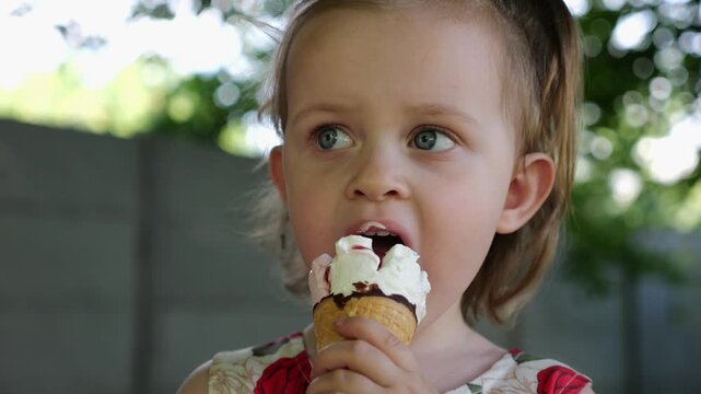 Portrait of attractive small girl eating delicious ice cream in a waffle cone outdoor. Beautiful child biting cold sweet dessert in summer garden. Cute kid enjoying tasty refreshing food on sunny day