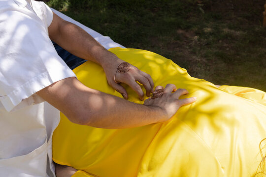 Therapist performing energy healing treatment on a person lying outdoors, hands gently placed on the back, wearing copper spiral bracelets, focusing on wellness and holistic health