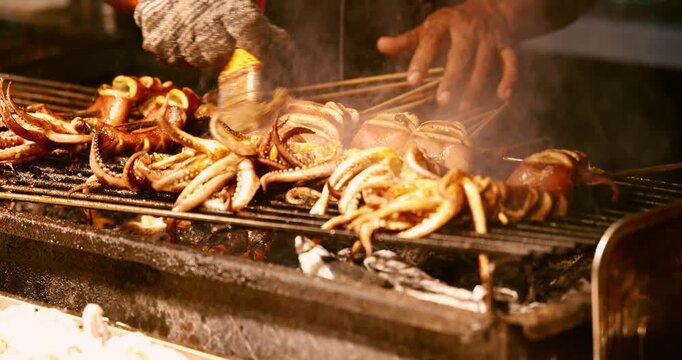 Grilled squid skewers brushed with sauce on charcoal grill at Asian night market, traditional street food preparation, culinary travel documentary.