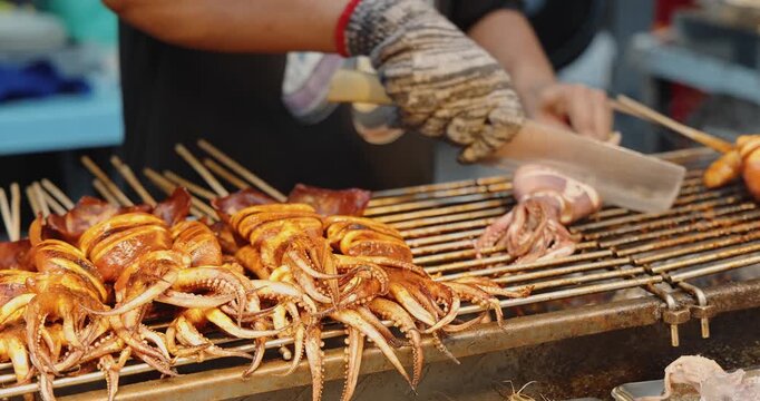 Grilled squid skewers being cut by vendor on outdoor grill, Asian street food market background, culinary travel documentary.