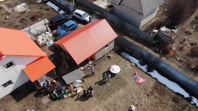 Aerial view of a rural house with orange roofs. Children play outside near a small garden. Snow patches linger beside a wooden fence. Scene suggests springtime family activity. Use for outdoor family