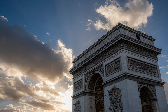 Arc de Triomphe in Paris France is framed by glowing clouds and backlit sky while the historic stone monument rises in dramatic evening light