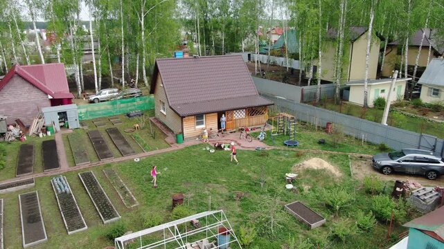 Aerial view of a rural garden with multiple raised beds. People tend to plots, walking among them with purpose. Greenhouse stands beside a parked car near a wooden house. Trees surround the yard