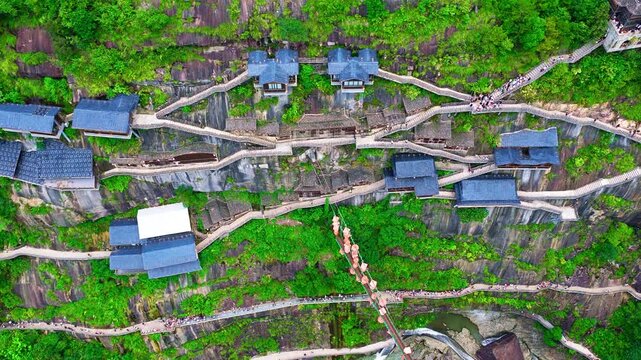 Aerial view of Wangxian Valley in Shangrao, Jiangxi, China, with winding stone stairways, blue-roofed pavilions, dense vegetation, and bright natural light.
