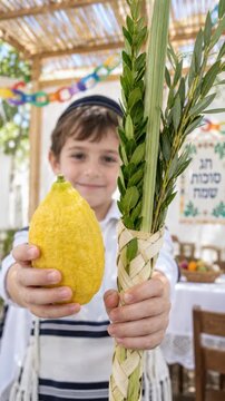 Jewish boy holding large etrog toward camera inside sukkah, lulav bundle beside smiling child in tallit, sukkot four species tradition for family harvest celebration