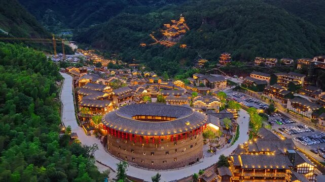 Aerial view of a forested mountain valley village with a large circular building, traditional rooftops, winding paths, and warm evening lights.