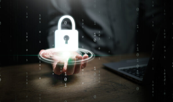 Cybersecurity concept. A man hand next to computer laptop in his office holding glowing padlock icon over working desk for system protection, security, and safety on dark background with copy space.