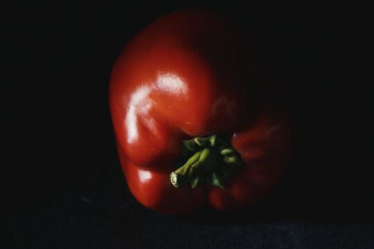 Close-up of ripe red bell pepper with vibrant green stem in moody lighting on dark background. Fresh raw vegetable for organic recipes, culinary menus, nutrition, and healthy eating.