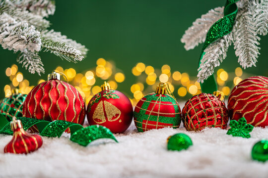 A Christmas background featuring an abundant array of red and green baubles with golden patterns beneath frosted pine branches, wrapped in ribbon on snow, set against deep green with lights.