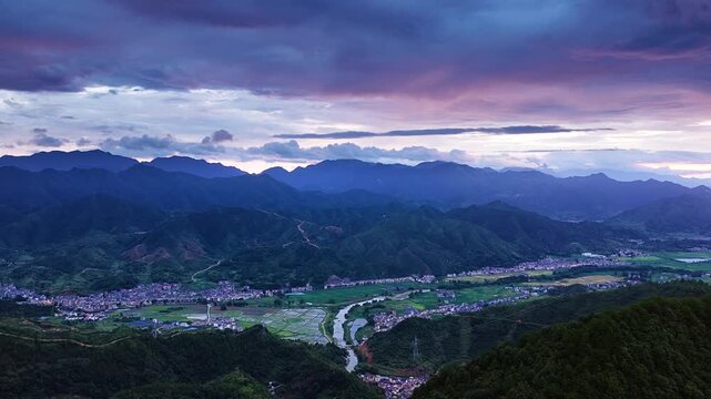 Mountain valley landscape with layered ridges, a winding river, patchwork fields, and clustered villages beneath blue-purple clouds and soft evening light.