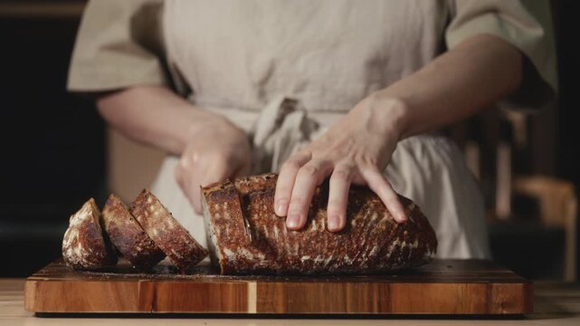 Close-up of baker in rustic linen apron slicing freshly baked artisan sourdough bread with knife on wooden cutting board. Homemade crusty loaf for breakfast, bakery promos, and healthy food concepts.