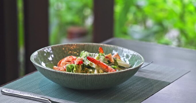Shrimp Caesar salad featuring grilled prawns and fresh lettuce in ceramic bowl. Served on table mat at outdoor restaurant. Natural daylight, blurred garden background, healthy dining.