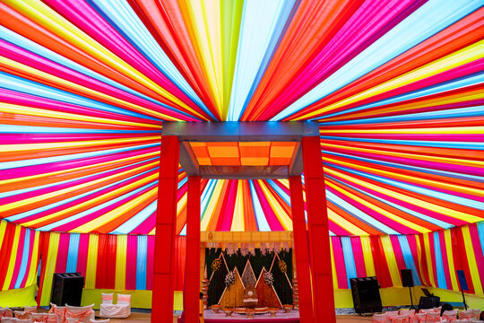 Colorful traditional Indian wedding event tent decorated with vibrant multicolored fabric drapes, featuring a beautifully decorated wedding stage and ceremonial mandapam setup.