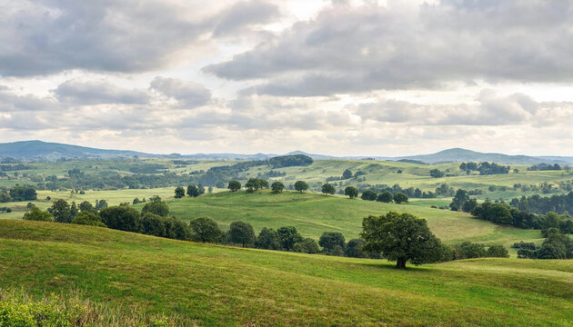 Rolling green hills under a cloudy sky, dotted with trees and farms, create a picturesque pastoral landscape.