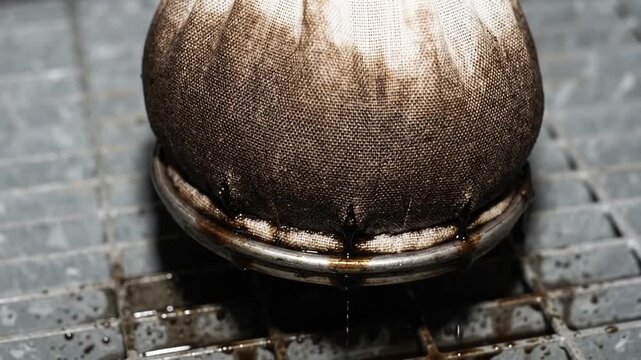 fabric filter bag dripping dark liquid through a metal strainer ring onto a steel grating floor