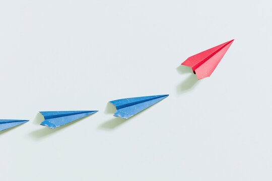 A single red paper plane breaking away from a row of blue planes, featuring a textured cardstock finish and soft shadows for business concepts and copy space