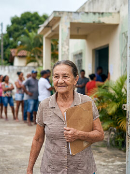Elderly woman holding a document folder outside a public clinic with people queuing in the background