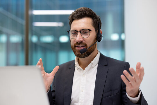 A close-up shot of a young businessman wearing a headset and a suit, sitting at a desk in an office, talking remotely and gesturing with his hands