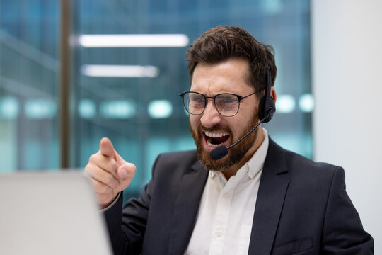 A close-up photo of an angry young businessman in a suit and headset in an office, speaking aggressively and pointing his finger at the laptop camera