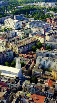 Vertical video. Geneva, Switzerland. Aerial sweep over the central city at morning light, dense historic rooftops and modern civic architecture unfolding beneath a clear sky. Drone footage. Rich colo
