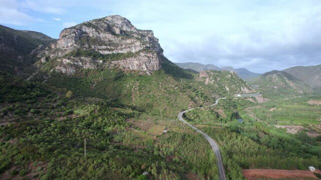 Stunning aerial view flying backwards from the majestic Monte Lora. A scenic road winds through the lush green valley in Sardinia, Italy