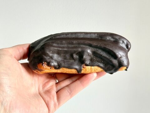 A person holds a delicious chocolate-glazed eclair in their hand against a neutral white background. A close-up food and pastry stock photograph captured indoors in Latvia for culinary projects.
