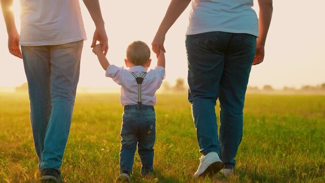 Parents, child walk hand in hand, Kid taking first steps, nature, sun. Mom dad son play outdoors. Happy family. Dad mom, kid boy taking walk in park, summer. Family picnic. Toddler learning to walk