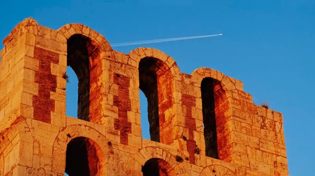 Odeon of Herodes Atticus stone arches in golden light with a jet trail in the sky in Athens, Greece