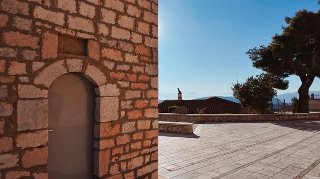 Sunlight shining brightly beside the Church of Agios Georgios with an arched doorway, overlooking a distant landscape in Arachova, Greece
